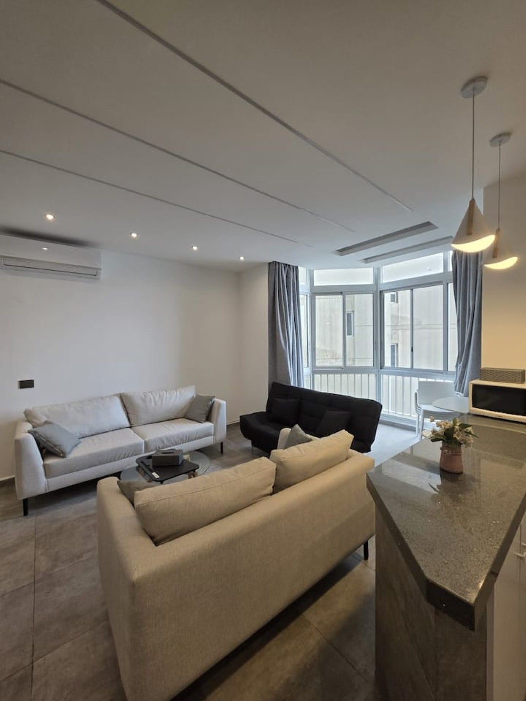 A modern living room with a sofa, a black armchair, and a coffee table. There is a large window with curtains, an air conditioning unit, and a pendant light.