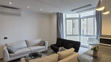 A modern living room with a sofa, a black armchair, and a coffee table. There is a large window with curtains, an air conditioning unit, and a pendant light.