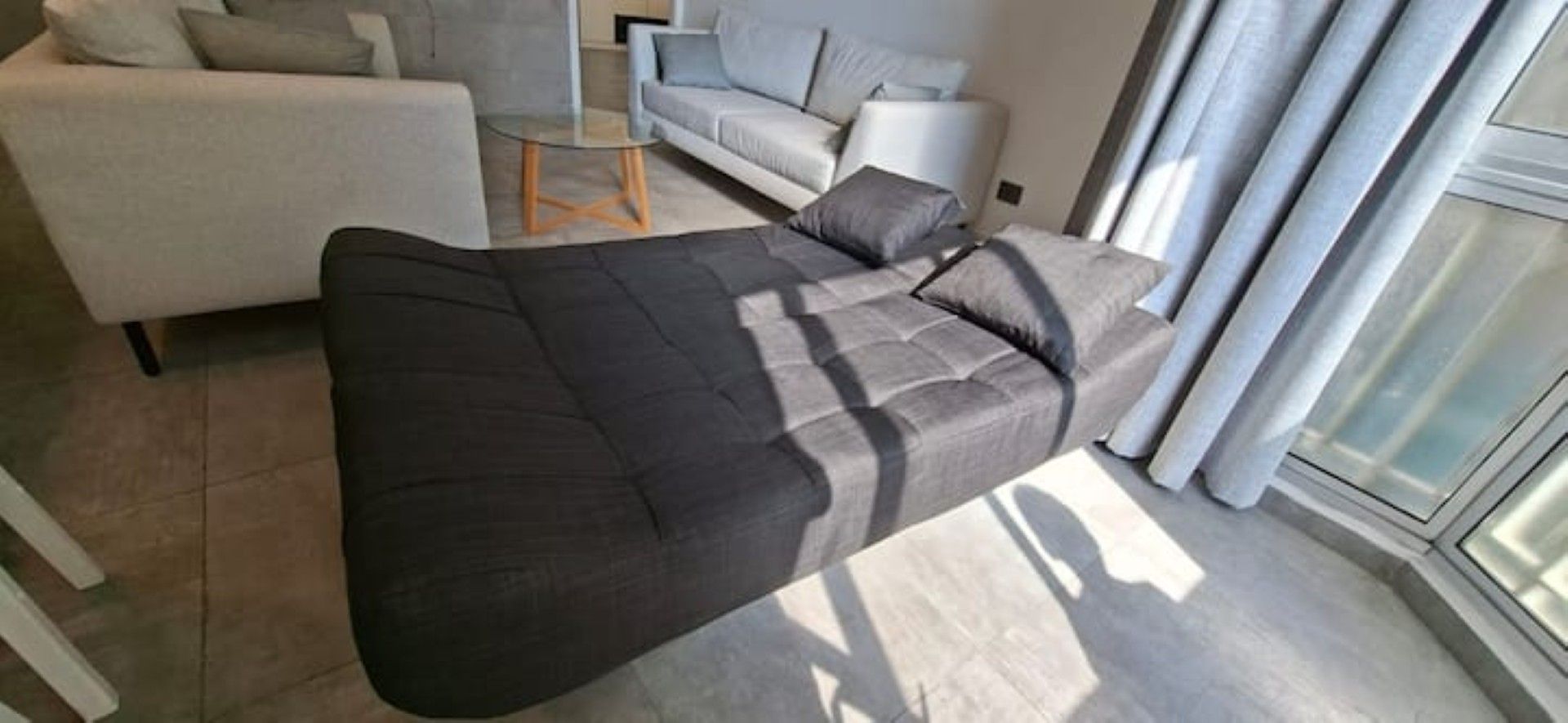 A modern living room with a dark sofa, a glass coffee table, and a light-colored sofa in the background.