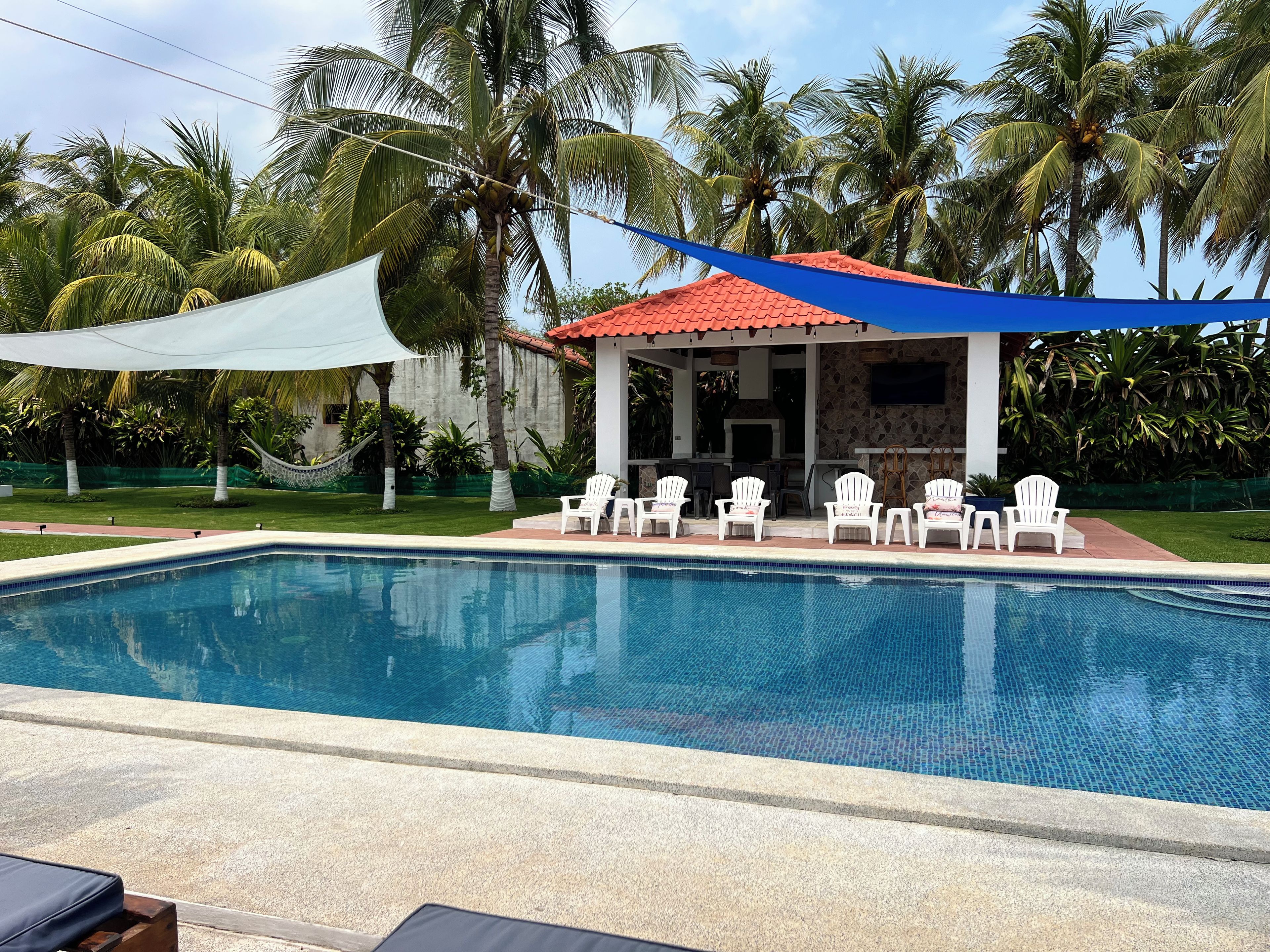 Ein Schwimmbecken mit Liegestühlen, einer überdachten Terrasse und Palmen im Hintergrund.