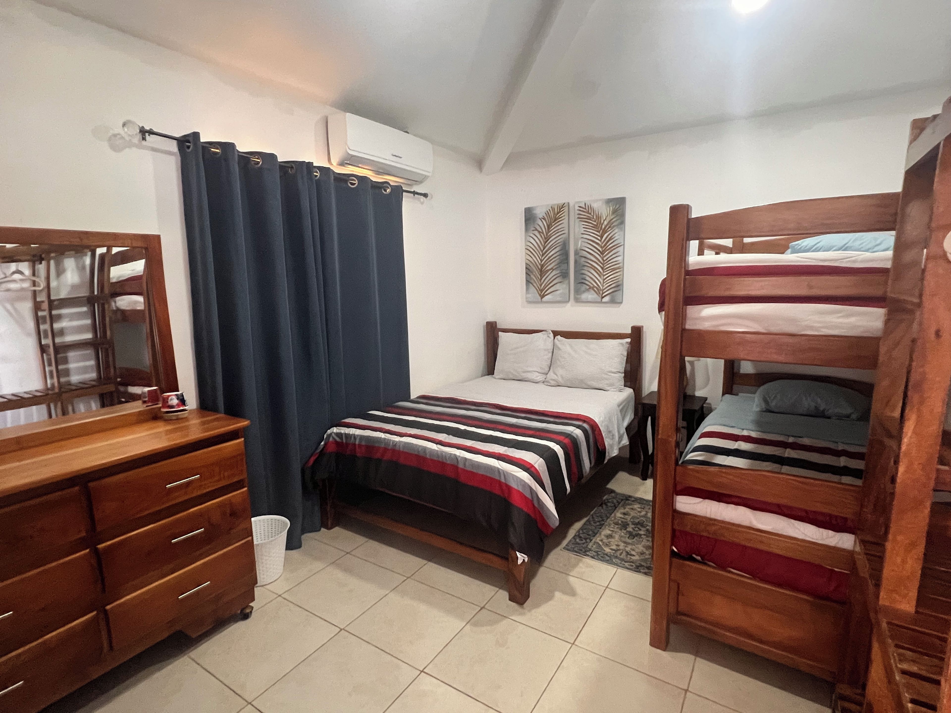 Une chambre à coucher comprenant un lit, une commode, un miroir, une fenêtre avec des rideaux et un appareil de climatisation.