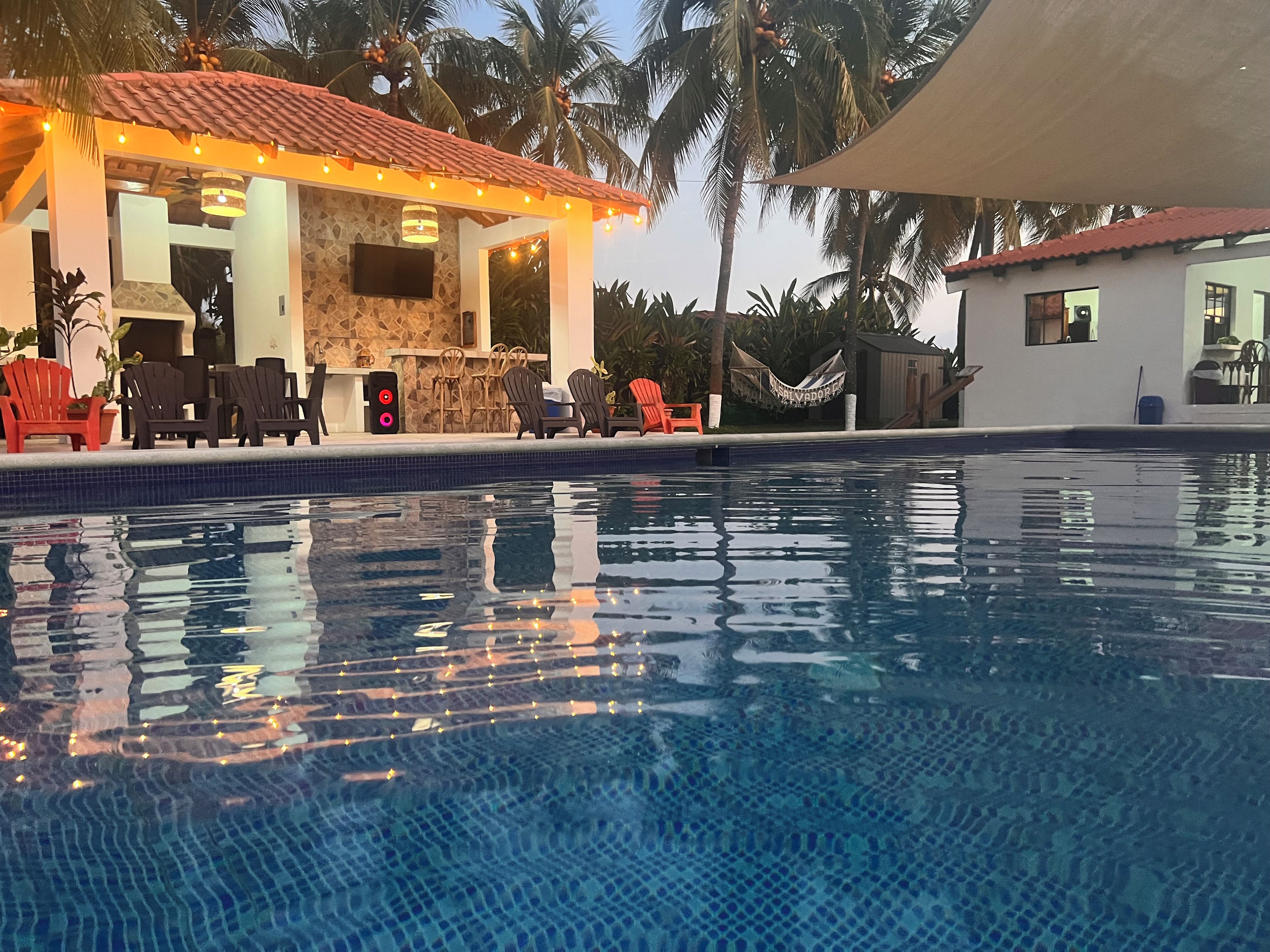 Une piscine avec un bord carrelé, un espace extérieur aménagé pour s’asseoir et un bâtiment doté d’une façade en pierre et d’un toit recouvert de tuiles rouges.