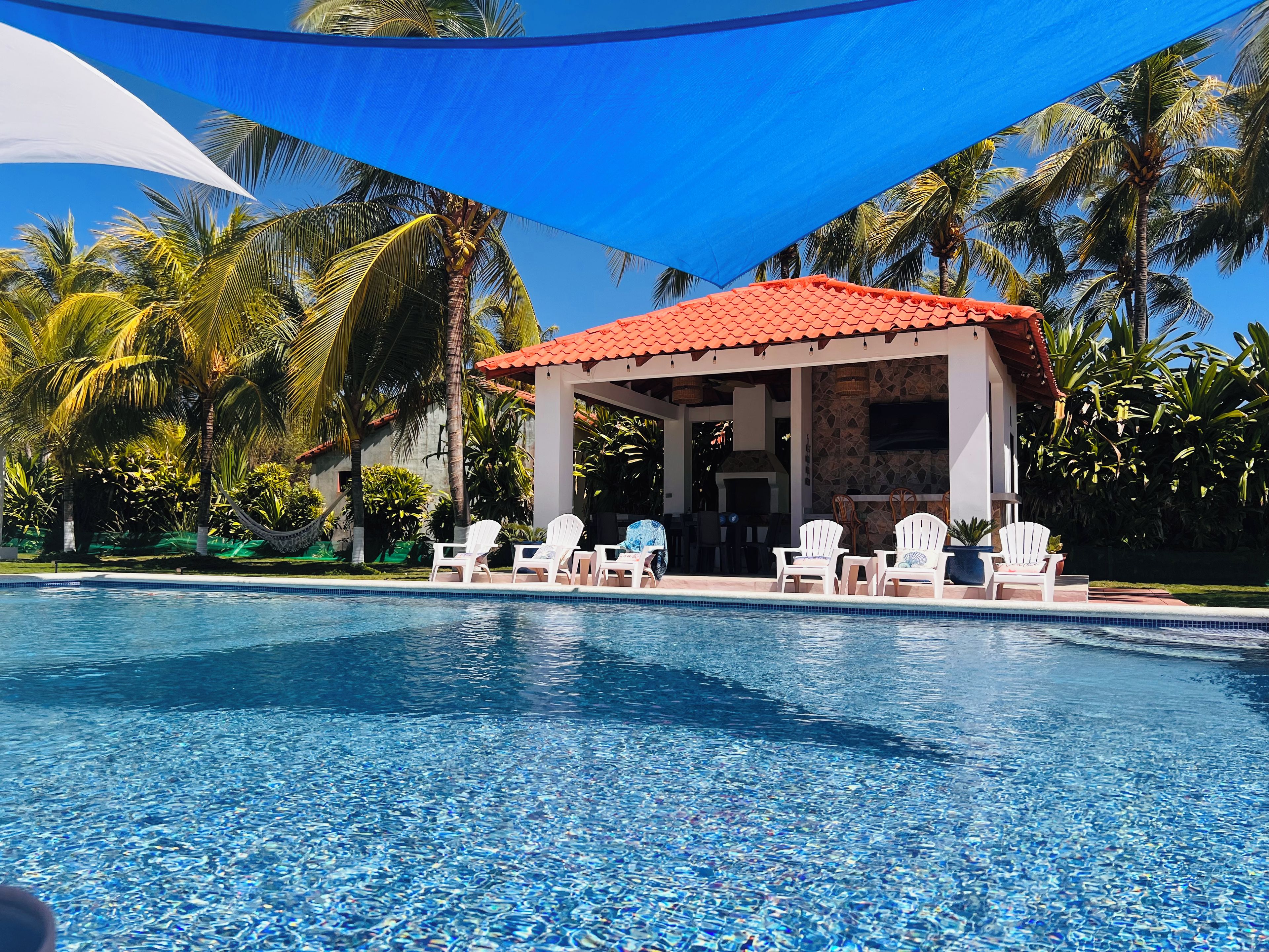 Area piscina con zona relax coperta, palme e piscina cristallina blu.