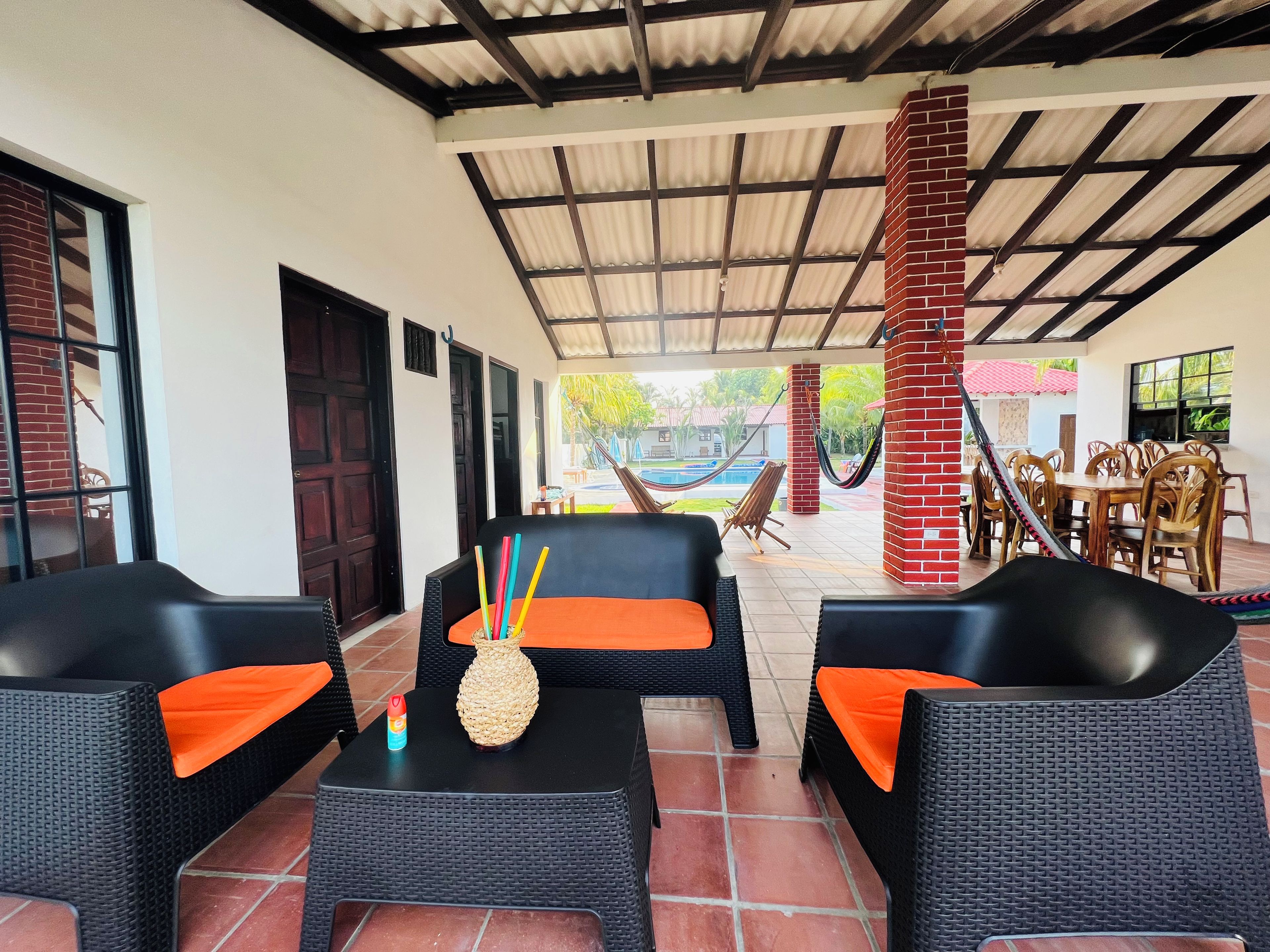 Ein Außenbereich mit Korbmöbeln und orangefarbenen Kissen, ein Pool im Hintergrund und eine überdachte Terrasse mit Metallveranda.