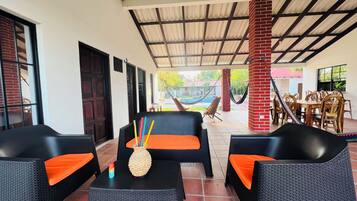 Ein Außenbereich mit Korbmöbeln und orangefarbenen Kissen, ein Pool im Hintergrund und eine überdachte Terrasse mit Metallveranda.