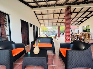 Ein Außenbereich mit Korbmöbeln und orangefarbenen Kissen, ein Pool im Hintergrund und eine überdachte Terrasse mit Metallveranda.