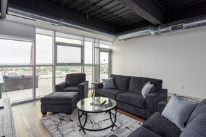 A modern living room with a grey sofa set, a glass coffee table, and a view of the city through large windows. - Porsche Suite (Buffalo)