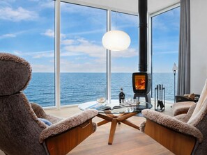 Quarto aconchegante com uma grande janela com vista para o mar, lareira a lenha, poltronas macias e uma mesa de vidro.