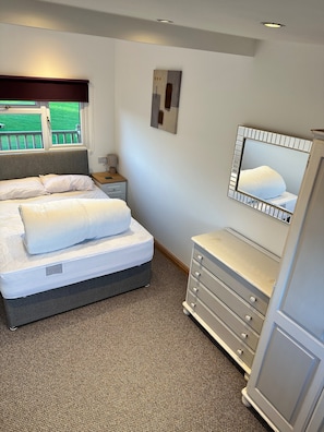 Un dormitorio con cama, mesita de noche, lámpara, cómoda, espejo y ventana con vistas.