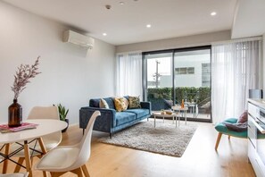 A modern living room with a sofa, a dining table, and chairs. There is a large window with curtains, a small table with a vase, and a plant. - Solara by Homello (Auckland)