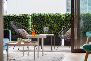 A modern outdoor seating area with round tables and chairs, a glass door leading to a balcony, and a green hedge wall. - Solara by Homello (Auckland)