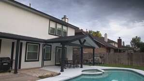 Un jardin avec une piscine, un patio couvert et un barbecue.