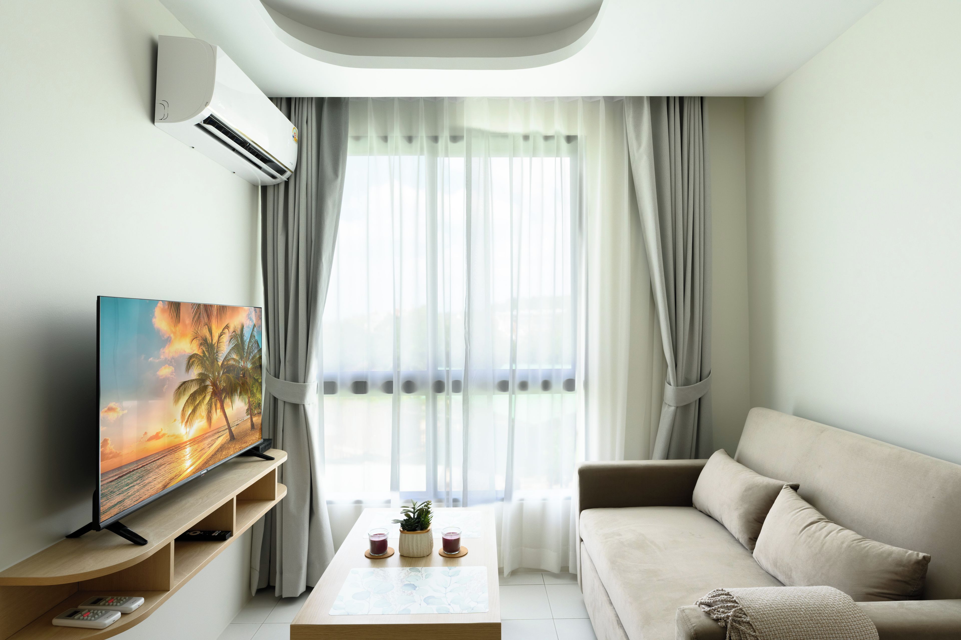 A modern living room with a flat-screen TV on a wooden stand, a beige sofa, a coffee table with two glasses of drink, and sheer curtains.