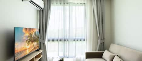 A modern living room with a flat-screen TV on a wooden stand, a beige sofa, a coffee table with two glasses of drink, and sheer curtains.