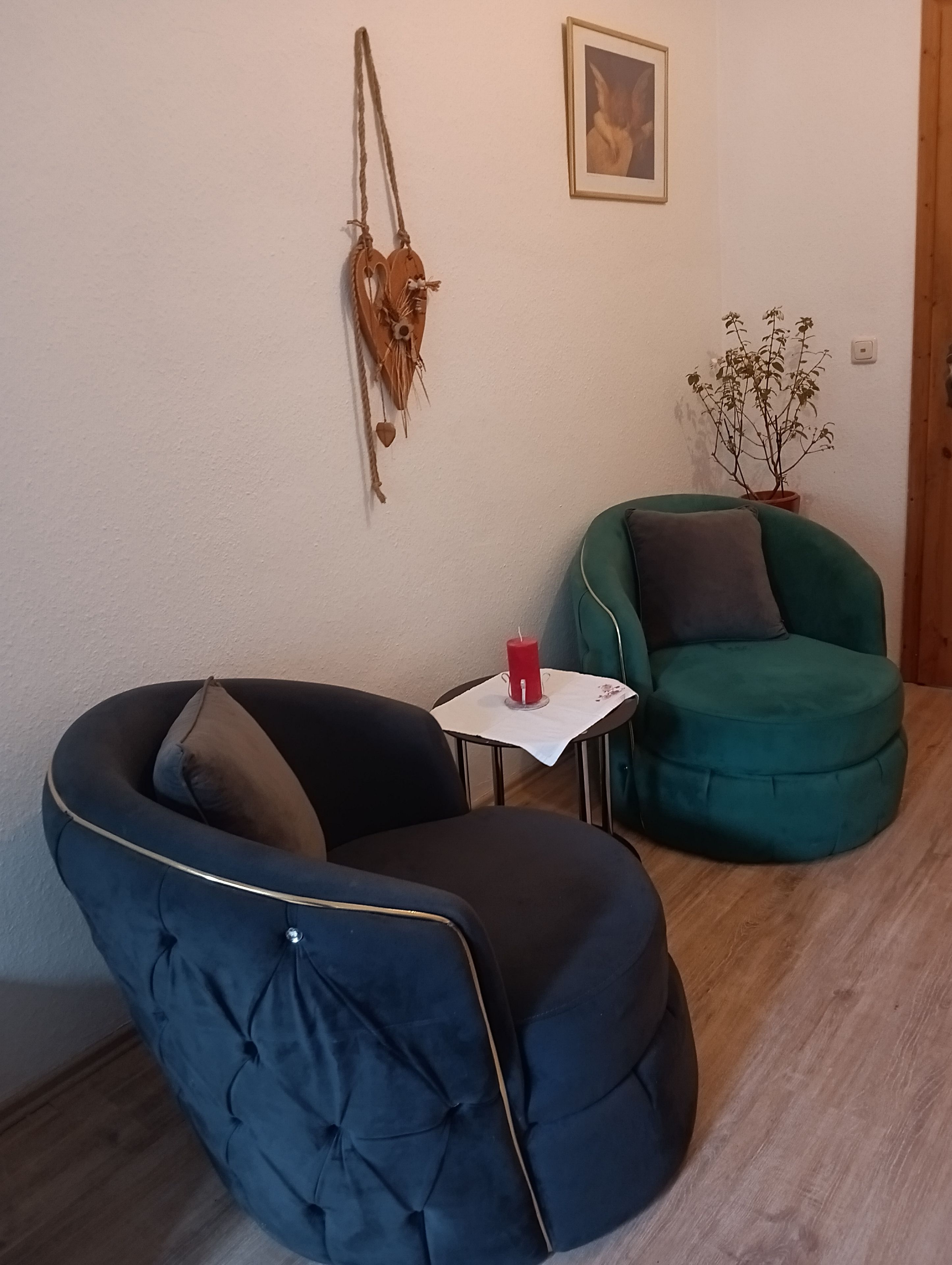 Ein Zimmer mit blauem Sessel, grünem Sessel, kleinem Tisch mit roter Kerze und Topfpflanze.