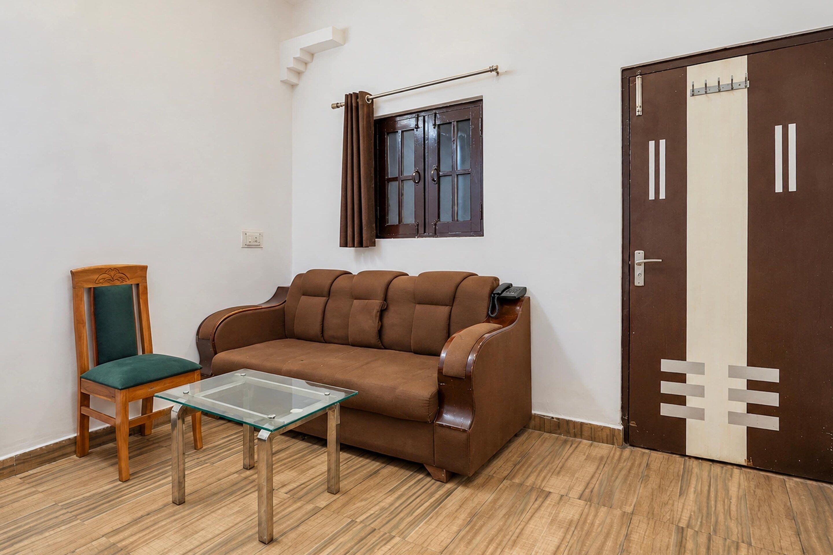 Una habitación pequeña con un sofá marrón, una silla de madera con cojín verde, una mesa con superficie de vidrio, una ventana con cortinas y una puerta con picaporte metálico.