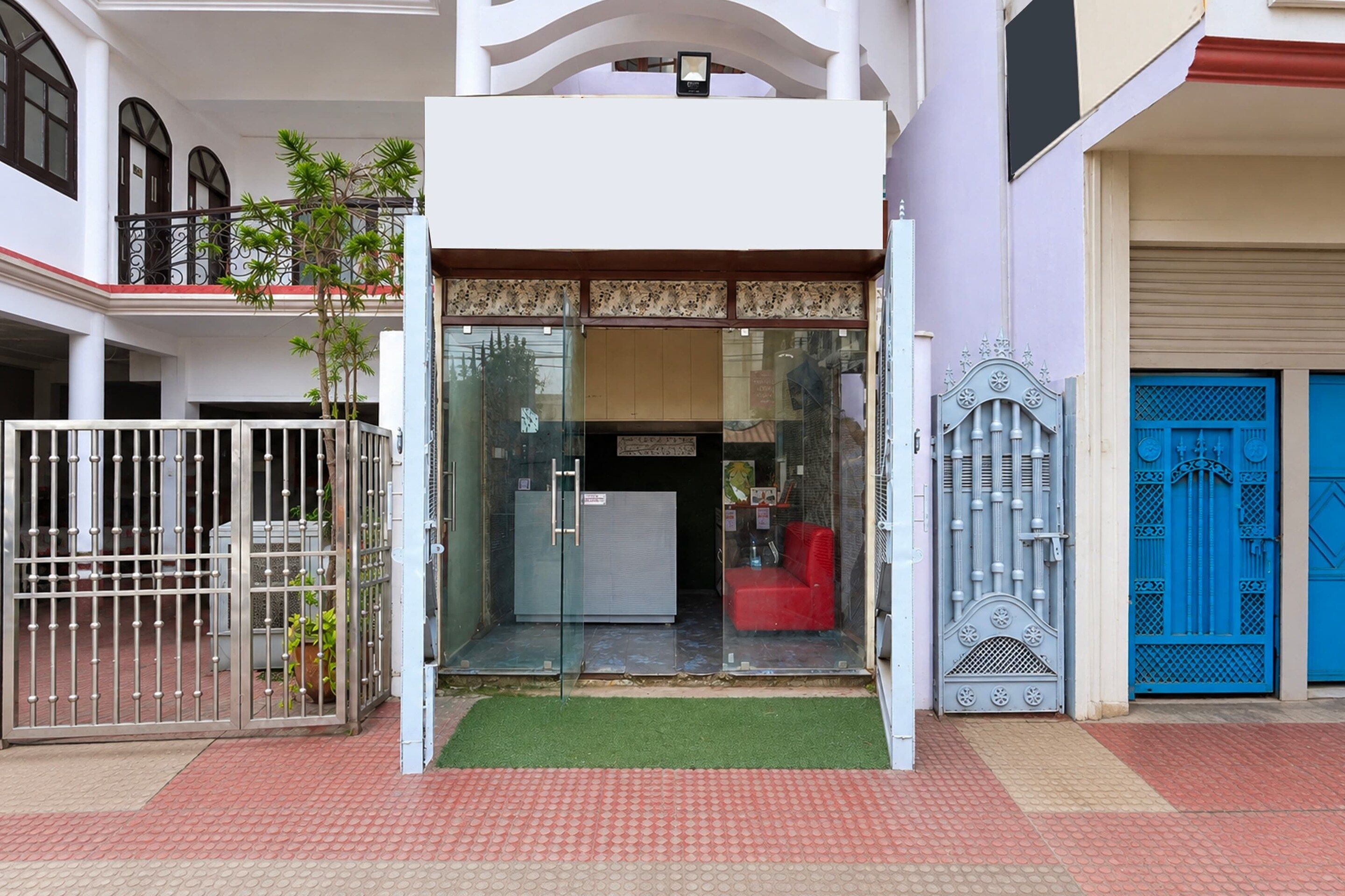 Una entrada de edificio con un camino de entrada cerrado por una reja, una pequeña zona de estar roja y una puerta azul.