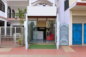 Una entrada de edificio con un camino de entrada cerrado por una reja, una pequeña zona de estar roja y una puerta azul.