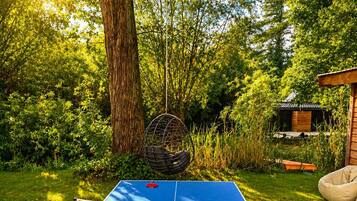 Een tafeltennistafel buiten, opgesteld in een tuin met een boom en een klein gebouw op de achtergrond.