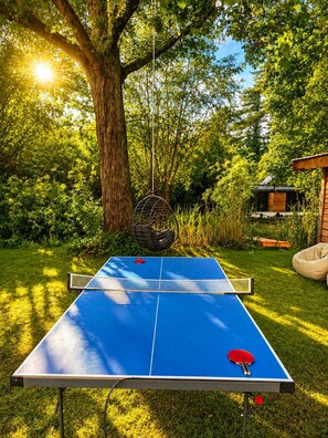 Een tafeltennistafel buiten, opgesteld in een tuin met een boom en een klein gebouw op de achtergrond.