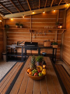 A wooden interior with a dining table centered, surrounded by wooden benches, a grill, and various kitchen utensils on the wall.