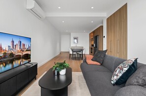 Ein modernes Wohnzimmer mit einem Flachbildfernseher, einem grauen Sofa und einer Essecke im Hintergrund.