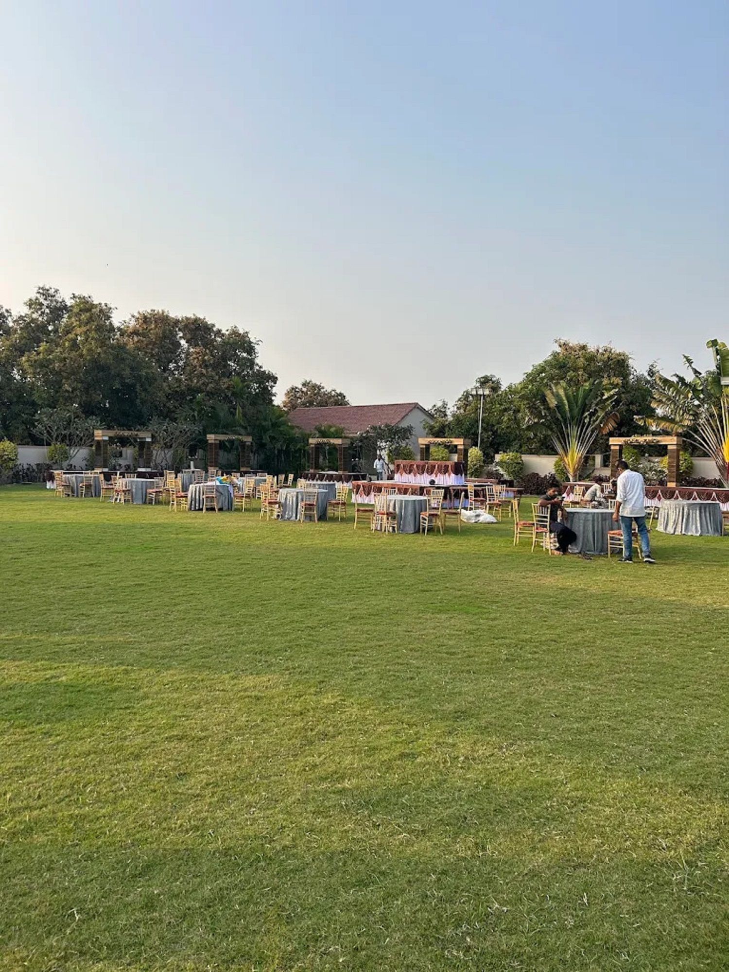 Un espacio al aire libre para eventos con varias mesas redondas y sillas, rodeado de césped y árboles.