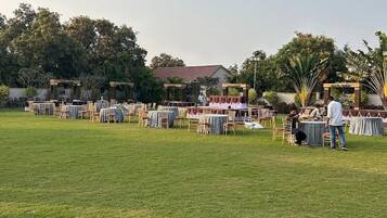 Un espacio al aire libre para eventos con varias mesas redondas y sillas, rodeado de césped y árboles.