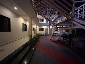 A corridor with a red and grey patterned floor, a staircase with a wooden railing, and a wall with a window and a door. - Rushi Resort By ARDEA (Valsad)