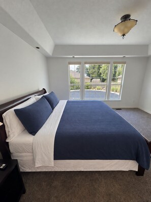 A bedroom with a large bed, a nightstand, a window with a view of the outdoors, and a ceiling light fixture. - Close to Lakeview Golf Course (Meridian)