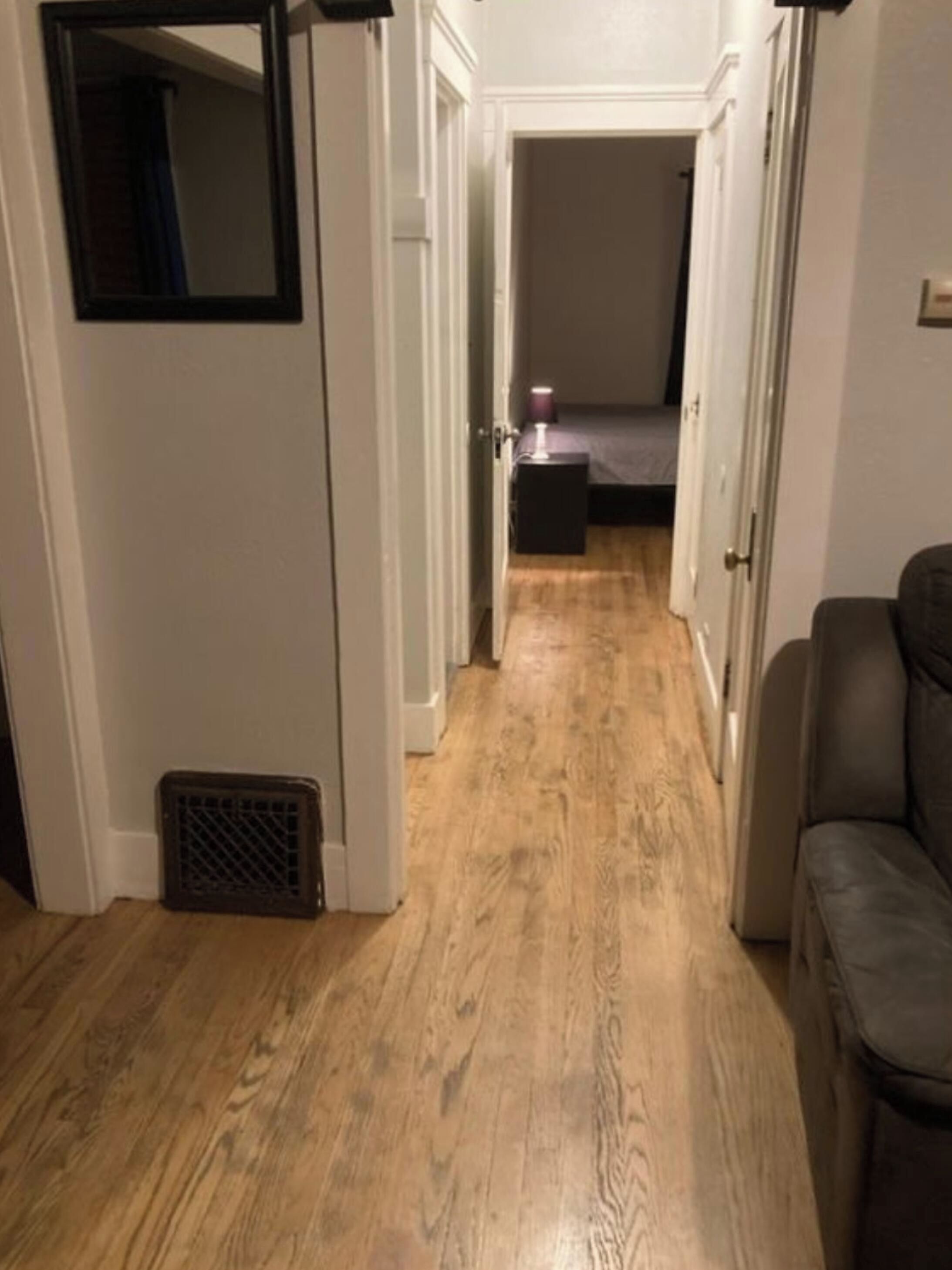 Um corredor estreito com piso de madeira, um respiradouro à esquerda e uma porta que dá para um quarto com cama e mesa de cabeceira.