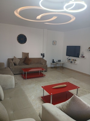 A modern living room with a sofa, a red coffee table, a flat-screen TV, and a ceiling light fixture.