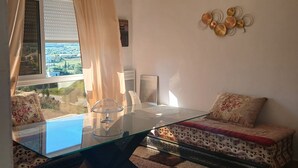 A modern room with a glass table, a sofa with patterned cushions, a decorative wall piece, and a view of a landscape through the window. - A lovely, beautiful view (Nefza)