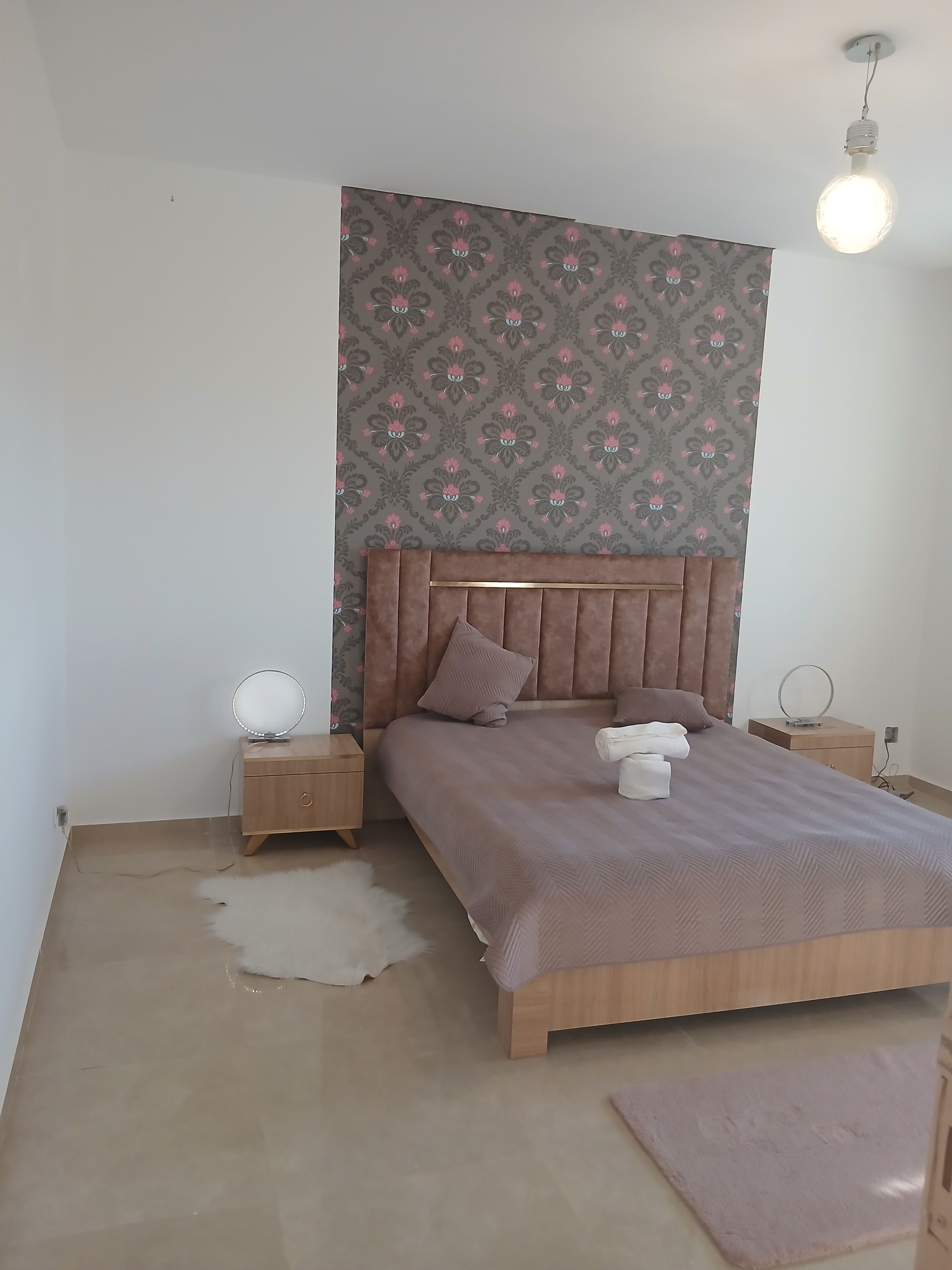 Une chambre avec un papier peint à motifs, une tête de lit en bois, une table de chevet ronde avec un miroir, un lit avec une tête de lit marron et un luminaire.