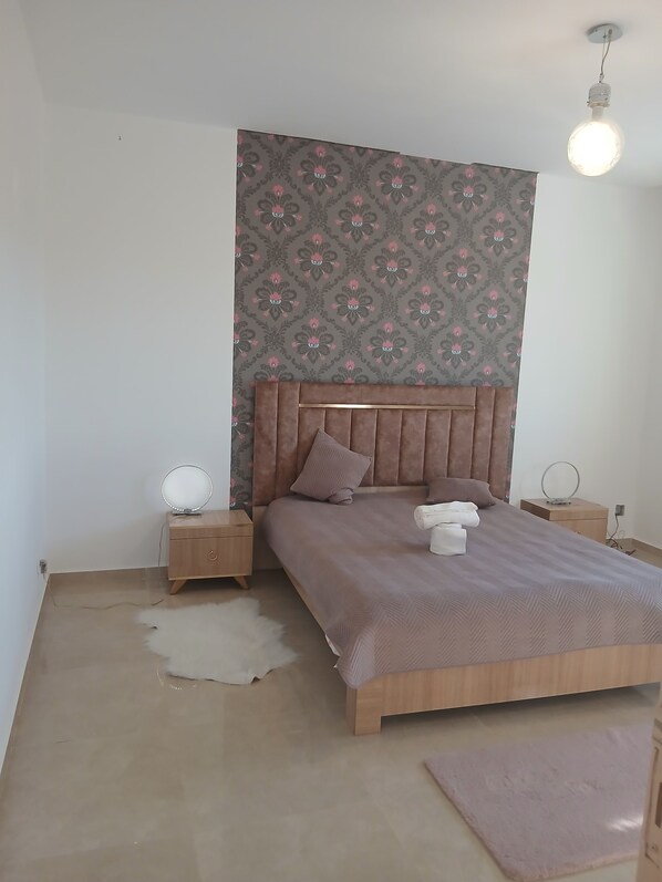 A bedroom with a patterned wallpaper, a wooden headboard, a round bedside table with a mirror, a bed with a brown headboard, and a light fixture.