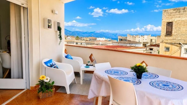 Un balcón con mobiliario blanco, una mesa con mantel estampado y vistas a edificios y montañas.