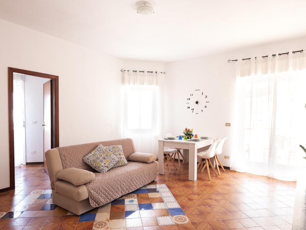 A living room with a sofa, a dining table with chairs, a wall clock, and a door.