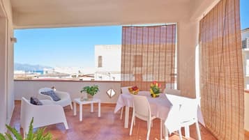 A balcony with white furniture, a table with a vase of flowers, and a view of buildings and mountains.