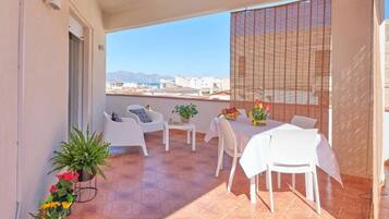 Un balcón con una mesa y sillas, plantas en macetas y vistas a edificios y montañas.