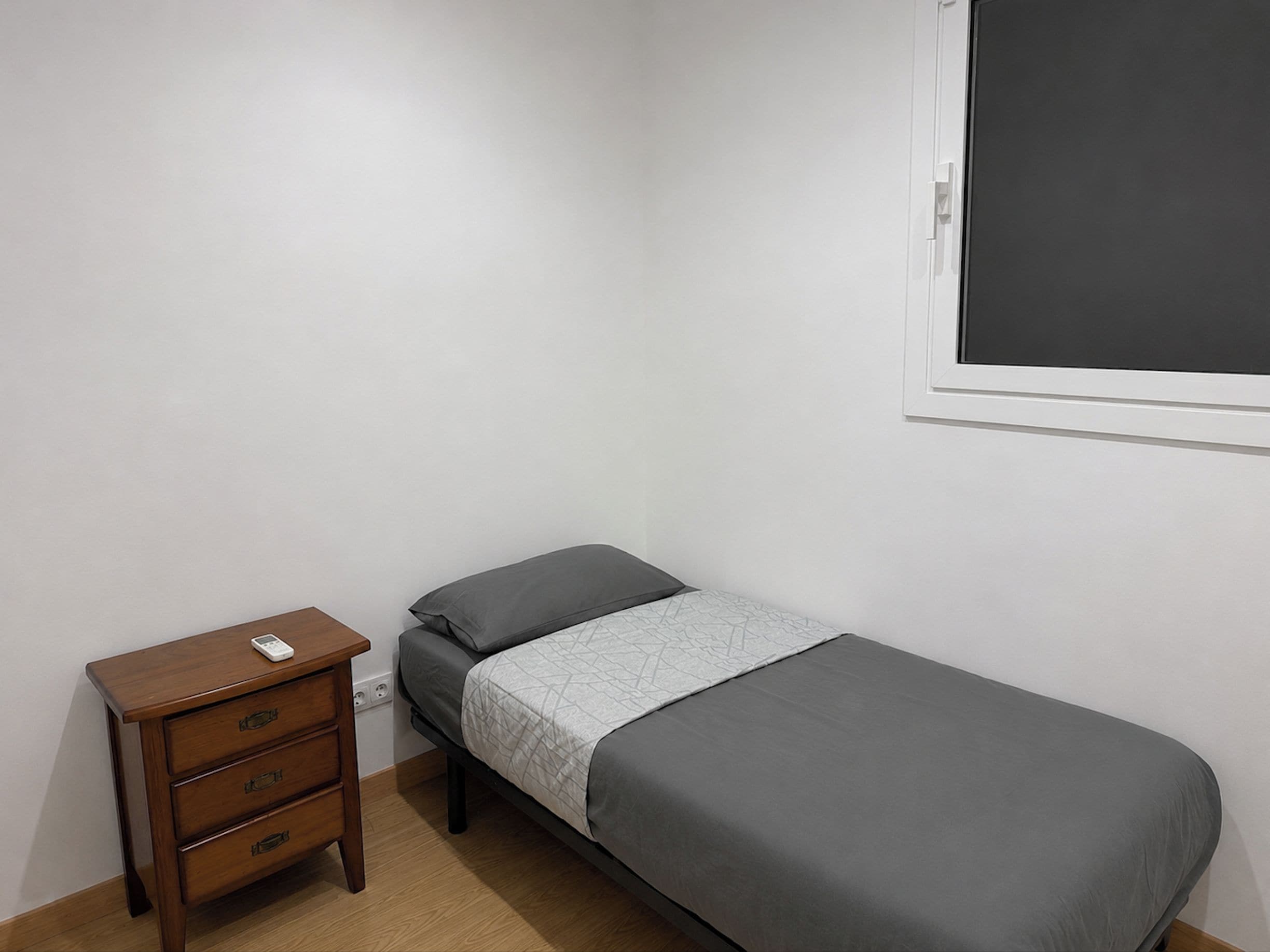 Een kleine kamer met een enkel bed, een houten nachtkastje, een grijze spreideken en een witte muur.