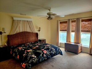 Une chambre à coucher comprenant un lit, une table de chevet, une lampe, un ventilateur de plafond et une fenêtre avec des stores.