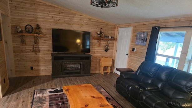 A cozy living room with a wooden floor, a black leather sofa, a wooden coffee table, a flat-screen TV, and a fireplace. - Trailside Lodge - Where Adventure Begins! (Shingleton)