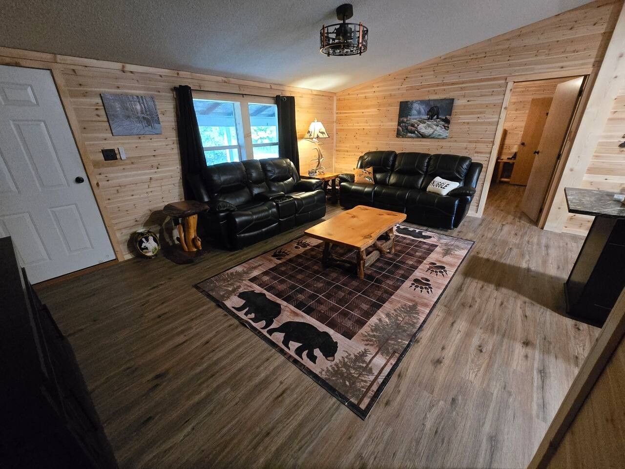 Un salon chaleureux avec un plancher en bois, deux canapés en cuir noir, une table basse en bois et un tapis orné d’un motif d’ours.