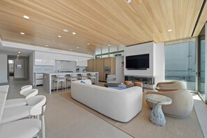 A modern living room with a large L-shaped sofa, a dining area with white chairs, a kitchen with white cabinets, and a large TV on the wall. - Newport Beach | Luxury Condo | Steps to Sand (Newport Beach)