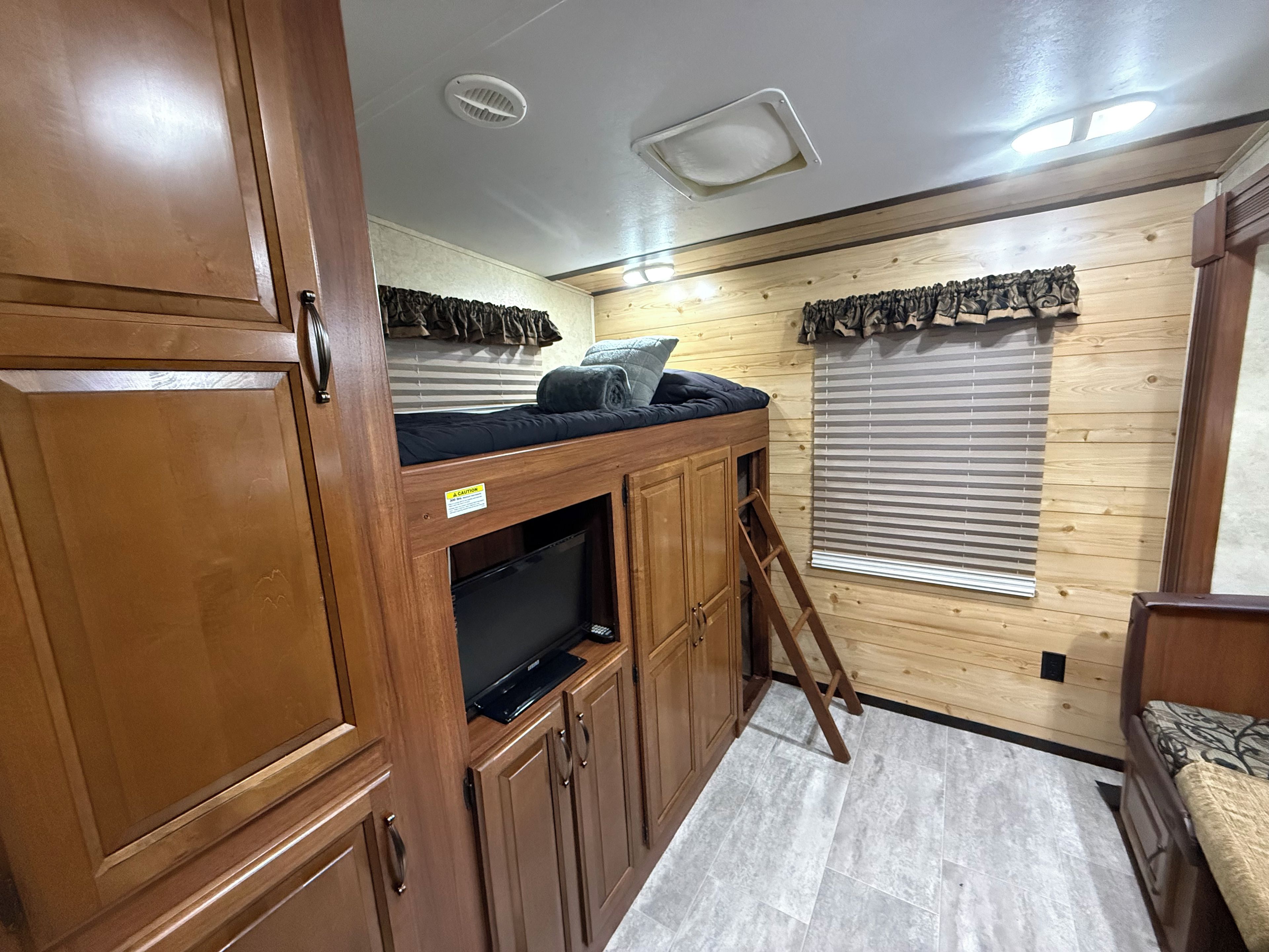 A compact RV interior with a kitchenette, a sleeping area with a mattress and pillows, and a small television on a cabinet.