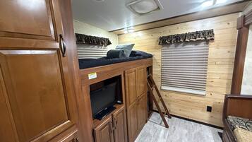 A compact RV interior with a kitchenette, a sleeping area with a mattress and pillows, and a small television on a cabinet.
