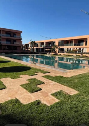 A swimming pool with a geometric pattern of grass and concrete, surrounded by a building with balconies and a clear blue sky. - Cozy 2-bedroom apartment in marvelous Marrakech with AC (Marrakech)