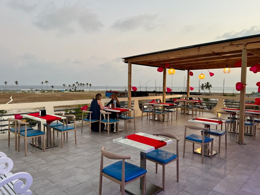 Een terras met tafels en stoelen, een overdekt zitgedeelte en uitzicht op het strand en de zee.