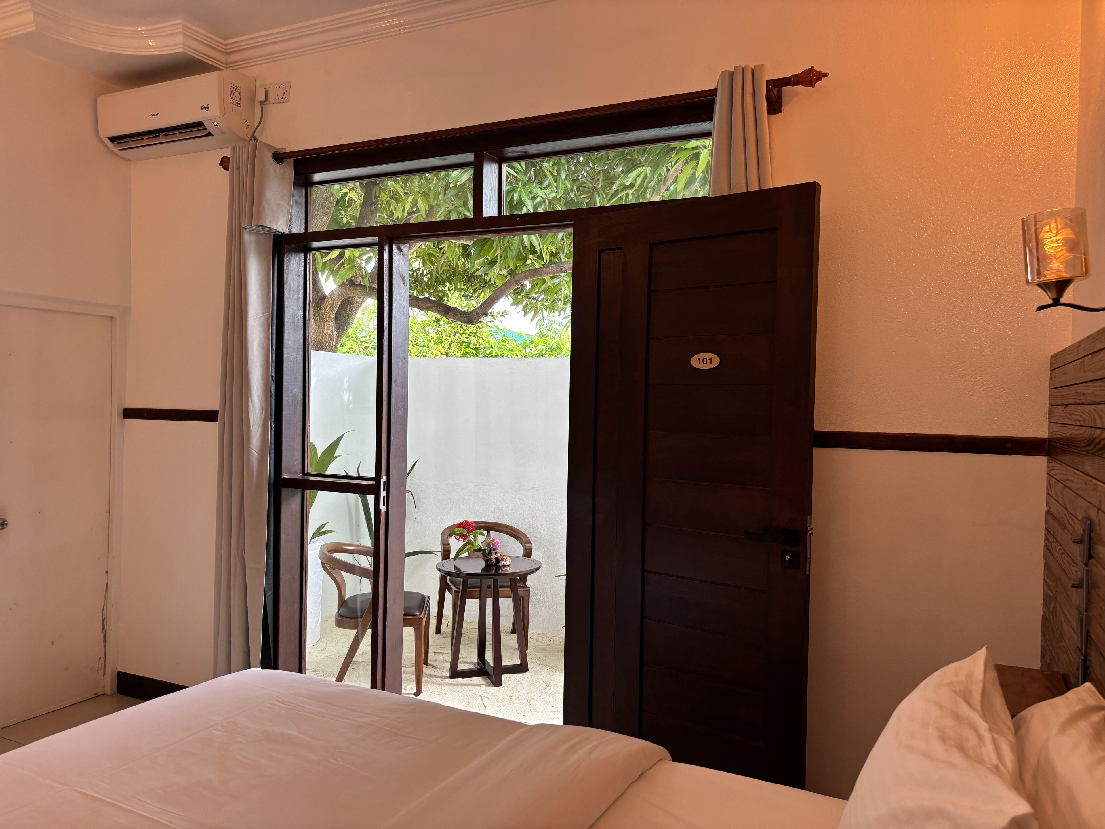 A hotel room with a bed, a small table with chairs, a view of greenery outside, and a door with a number.