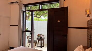 A hotel room with a bed, a small table with chairs, a view of greenery outside, and a door with a number.